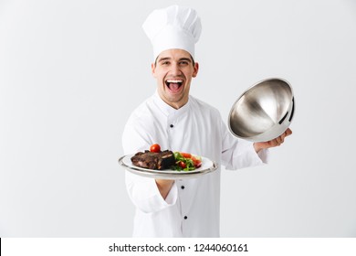 Un hombre muy emocionado cocinero que usa un velo de vestir uniforme aislado sobre fondo blanco, mostrando un plato de carne