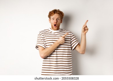 Excited Man With Curly Red Hair Showing Promo Offer And Looking Amazed, Pointing Fingers At Upper Right Corner Logo, White Background