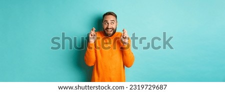 Excited man expecting something with anticipation, hold fingers crossed and smiling, making wish, standing over light blue background.