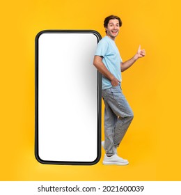 Excited Man Leaning On Big Smartphone With Blank White Screen And Gesturing Thumb Up Sign, Cheerful Guy Recommending New App Or Website, Standing On Yellow Background, Mock Up Image, Full Body Length