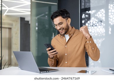 Excited man in a modern office celebrating success by looking at his smartphone and raising his fist in joy. - Powered by Shutterstock