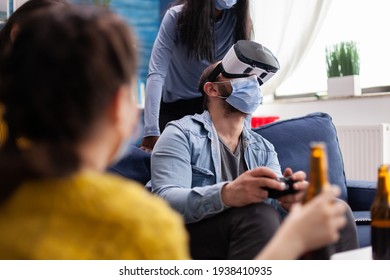 Excited man spending time with friends experiencing virtual reality playing games with vr headset wearing face mask to prevent coronavirus spread in global pandemic. Diverse people having fun at new