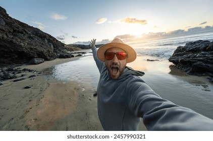 Excited man in straw hat and sunglasses taking a selfie on a sandy beach at sunset and enjoying summer holidays  - Powered by Shutterstock