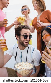 Excited Man In Sunglasses Toasting With Beer Near Asian Woman And Blurred Multiethnic Friends