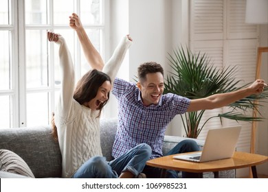 Excited man and woman screaming with joy raising hands looking at laptop screen sitting on sofa at home, happy young couple celebrate online win victory, goal achievement, good news, new opportunity