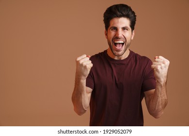 Excited masculine man screaming while making winner gesture isolated over beige background
