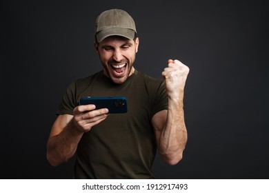 Excited Masculine Military Man Playing Online Game On Mobile Phone Isolated Over Black Background