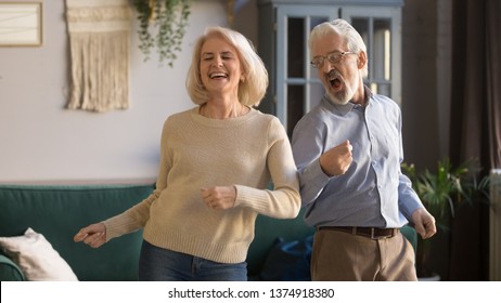 Excited mature couple, laughing grey haired man and woman having fun, dancing to favorite music at home in living room, middle aged wife and husband spending weekend together, funny activity