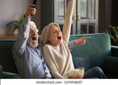 Excited Mature Couple, Middle Aged Man And Woman Watching Football On Tv, Senior Sport Fans Celebrating Favorite Team Victory, Sitting On Cozy Couch And Eating Popcorn Snack At Home, Enjoying Weekend