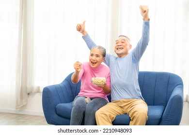 Excited Mature Couple, Senior Man And Woman Watching Tv, Senior Sport Fans Celebrating Favorite Team Victory, Sitting On Cozy Couch And Eating Popcorn Snack At Home, Enjoying Weekend. Happy Senior.