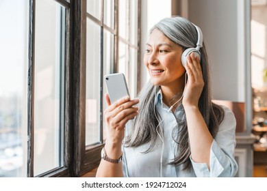 Excited mature grey-haired woman listening to the music on the phone at home - Powered by Shutterstock