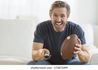 Excited Mature Man With American Football Watching TV On Sofa