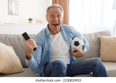 Excited Mature Man Watching Football Match, Shaking Clenched Fist. Emotional Guy Sitting On Couch Cheering Favorite Team Enjoying Game Goal On TV Holding Remote Controller And Ball, Looking At Camera