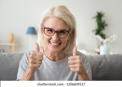Excited mature woman in glasses sit on couch at home showing thumbs up satisfied with service, smiling elderly female make like gesture recommending something, satisfied with choice or decision