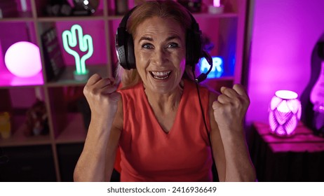 Excited mature woman with headphones in neon-lit gaming room celebrating victory at night. - Powered by Shutterstock