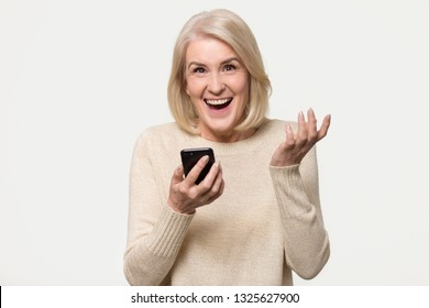 Excited mature woman looking at camera holding phone amazed by mobile win, happy overjoyed middle aged old lady winner surprised by great news in sms message isolated on white grey studio background