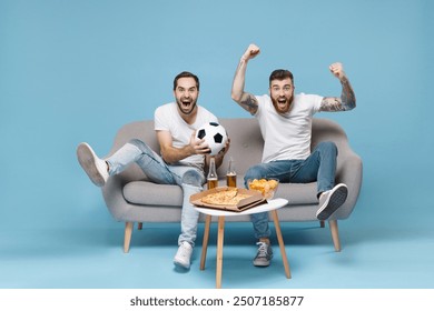 Excited men guys friends in white t-shirt sit on couch isolated on blue background. Sport leisure concept. Cheer up support favorite team with soccer ball scream expressive gesticulating with hands - Powered by Shutterstock