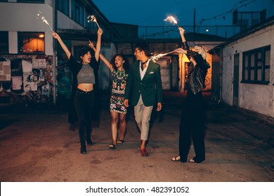 Excited Men And Woman Walking On City Street At Night Holding Sparklers. Group Of Friends Enjoying With Sparklers In Evening.