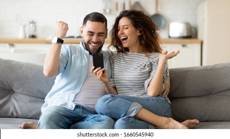 Excited millennial husband and wife relax on couch in kitchen feel euphoric win lottery online on cellphone, overjoyed young couple triumph celebrate read good unexpected news on smartphone gadget