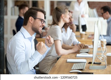 Excited millennial male employee work in shared workplace winning online lottery, happy worker gesture yes getting job promotion achieving goal, satisfied man feel euphoric reading great news online