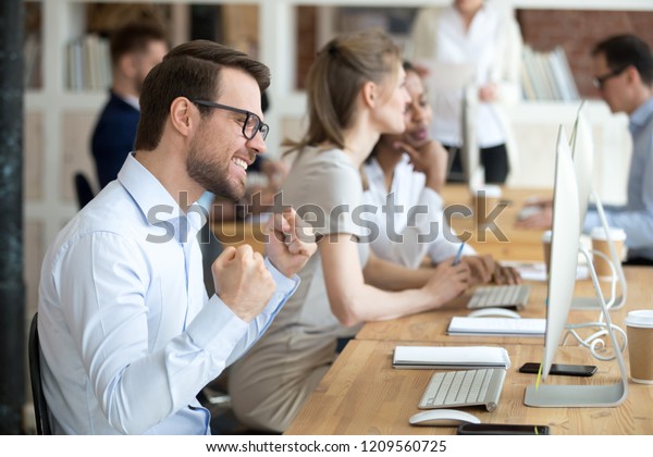Excited millennial male employee work in shared workplace winning online lottery, happy worker gesture yes getting job promotion achieving goal, satisfied man feel euphoric reading great news online