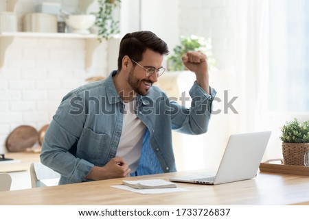 Excited millennial man in glasses sit at table in kitchen feel euphoric win online lottery on laptop, overjoyed young male in spectacles look at computer screen triumph get good news on email