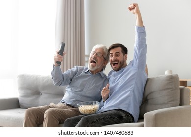 Excited millennial man and senior father sit on couch watching football game on TV raise hands cheering, happy elderly dad and young son relax on sofa support team enjoy match at home together - Powered by Shutterstock