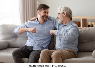 Excited millennial man sit on couch spend time with elderly father talking enjoying leisure time at home, smiling grown son relax on sofa with senior dad give fists bump, spend family weekend together