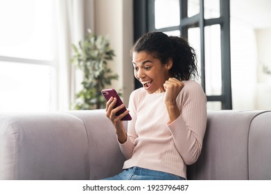 Excited Mixed-race Teenage Girl Celebrating Success, Winner Of The Competition Showing Hand Fist And Looking At Mobile Phone, Sitting On The Couch And Screaming Yes, Passed The Test, Achieved Goal