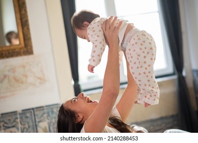 Excited Mother Tossing Her Baby Son In Bedroom. Portrait Of Young Happy Woman Holding Her Son At Home. Motherhood Concept