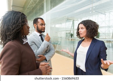 Excited multiethnic colleagues arguing outside. Business man and women standing at outdoor glass wall, talking to each other, gesturing. Work issues concept