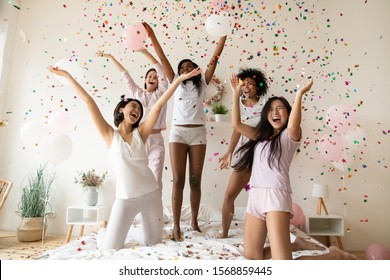 Excited Multiethnic Young Women Friends Wear Pyjamas Dance On Bed Together In Falling Confetti Having Fun Celebrate Bachelorette Bridal Shower, Happy Ladies Jump Enjoy Pajama Birthday Party Concept