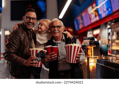 An excited multigenerational family is at the movie theater. Grandfather and his son are holding his daughter and beverages and popcorn in their hands looking at the camera. - Shutterstock ID 2274424021