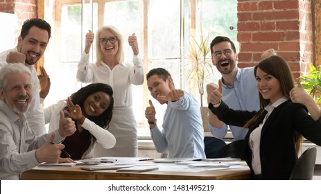 Excited multiracial businesspeople sit at office desk at meeting look at camera showing thumbs up together, overjoyed successful diverse colleagues posing for picture feel motivated at briefing