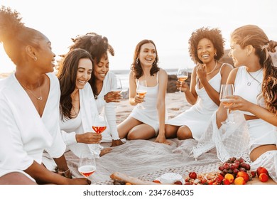 Excited multiracial female friends chatting, enjoying summer picnic with wine or champagne at sunset on beach, ladies having hen party on coastline - Powered by Shutterstock