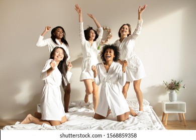 Excited Multiracial Girls Friends Bride And Bridesmaid Having Fun Dancing On Bed Together, Happy Diverse Young Ladies Wear Robes Laughing Jumping In Bedroom Celebrate Hen Bachelorette Party Concept