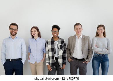 Excited Multiracial Millennial Team Standing Near Wall Looking At Camera Laughing, Group Of Young Diverse Employees Or Professionals Posing, Smiling Business People Or Staff Make Picture In Office