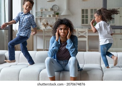 Excited naughty, noisy African children running around mom on sofa, shouting, making mother upset, annoyed. Frustrated mum sitting on couch at home, feeling headache, stress. Parenting problems