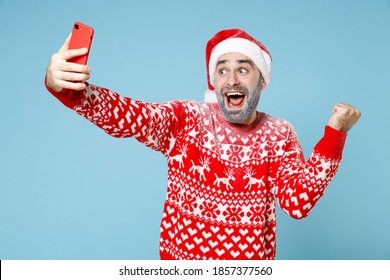 Excited Northern Bearded Man Frozen Face In Santa Hat Christmas Sweater Doing Selfie Shot On Mobile Phone Doing Winner Gesture Isolated On Blue Background. Happy New Year Holiday Winter Time Concept