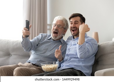 Excited old father and son watching tv, eating popcorn snack, having fun at home, family fans watching football or soccer match together, celebrating favorite team win, goal, sitting on couch