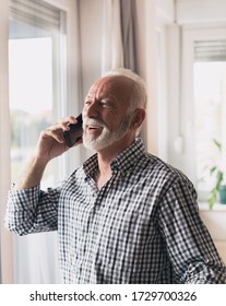 Excited Old Man In Shirt With White Hair And Beard Talking On Phone Beside Window At Home