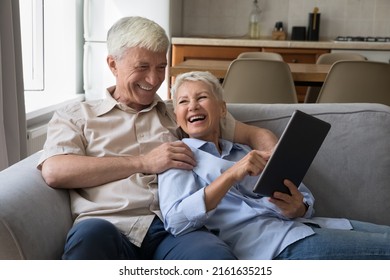 Excited older age grandparents family couple hug on couch at home use touchpad laugh watching funny video enjoy online shopping. Happy senior husband wife having fun with pad pc discuss web purchase