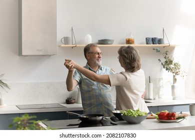 Excited Older Married Couple Celebrating Anniversary, Dancing To Music In Home Kitchen At Table With Fresh Vegetables Ingredients, Preparing Family Dinner Together, Enjoying Healthy Active Lifestyle