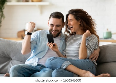 Excited overjoyed couple resting on couch holding smart phone celebrating on-line lottery win, bid betting victory moment, unbelievable opportunity or invitation, internet sale, getting prize concept