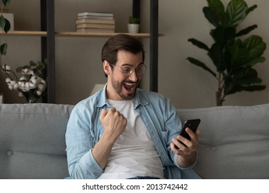 Excited overjoyed young man sit on sofa holding smartphone feeling euphoric with mobile online bet gambling victory. Ecstatic guy looks at cellphone scream with joy celebrate fantastic news received