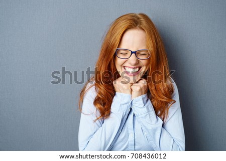 Excited overjoyed young woman wearing glasses beaming with happiness and screwing up her eyes as she holds her fists to the over a blue studio background with copy space