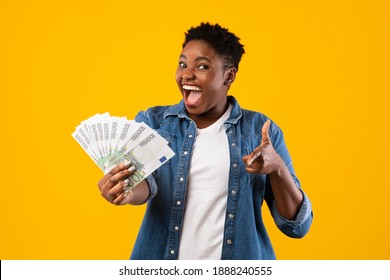 Excited Overweight African Woman Holding European Money Cash Pointing Finger At Camera Standing Over Yellow Background. Lady Posing With Euro Banknotes. Financial Success, Profit And Currency Concept