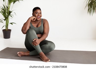 Excited overweight young black woman in tight sportswear sitting on yoga mat and smiling at camera, practicing yoga at home in the morning, panorama with copy space. Healthy lifestyle concept
