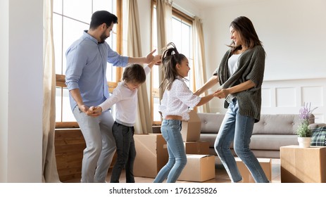 Excited Parents With Children Celebrating Moving Day In New House, Happy Mother And Father Dancing To Music With Adorable Little Son And Daughter In Living Room With Cardboard Boxes