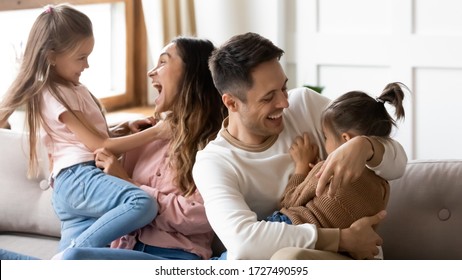 Excited parents having fun with two little daughters at home, cute preschool girls with loving happy mother and father sitting on couch at home, hugging and laughing, family spending weekend together